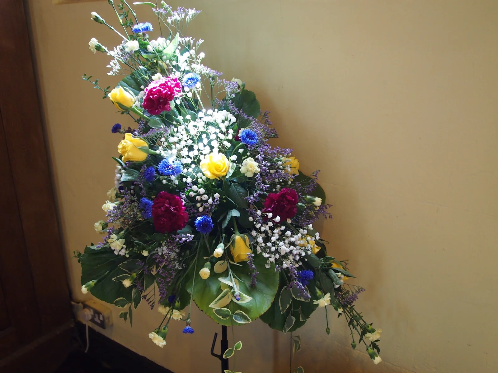 A floral arrangement of yellow roses, pink carnations and blue cornflowers against a wall.