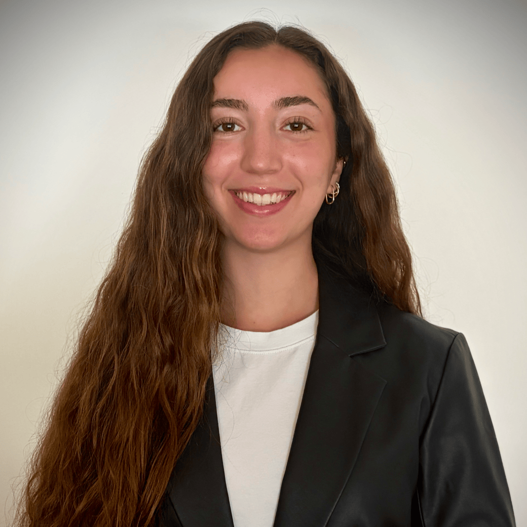 Young woman with long wavy brown hair wearing a black blazer and white shirt, smiling at the camera against a light background.