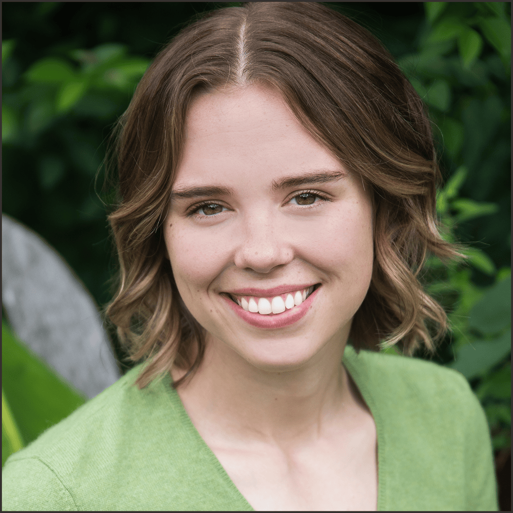 Smiling young woman with short brown hair in a green sweater, outdoors with greenery in the background.