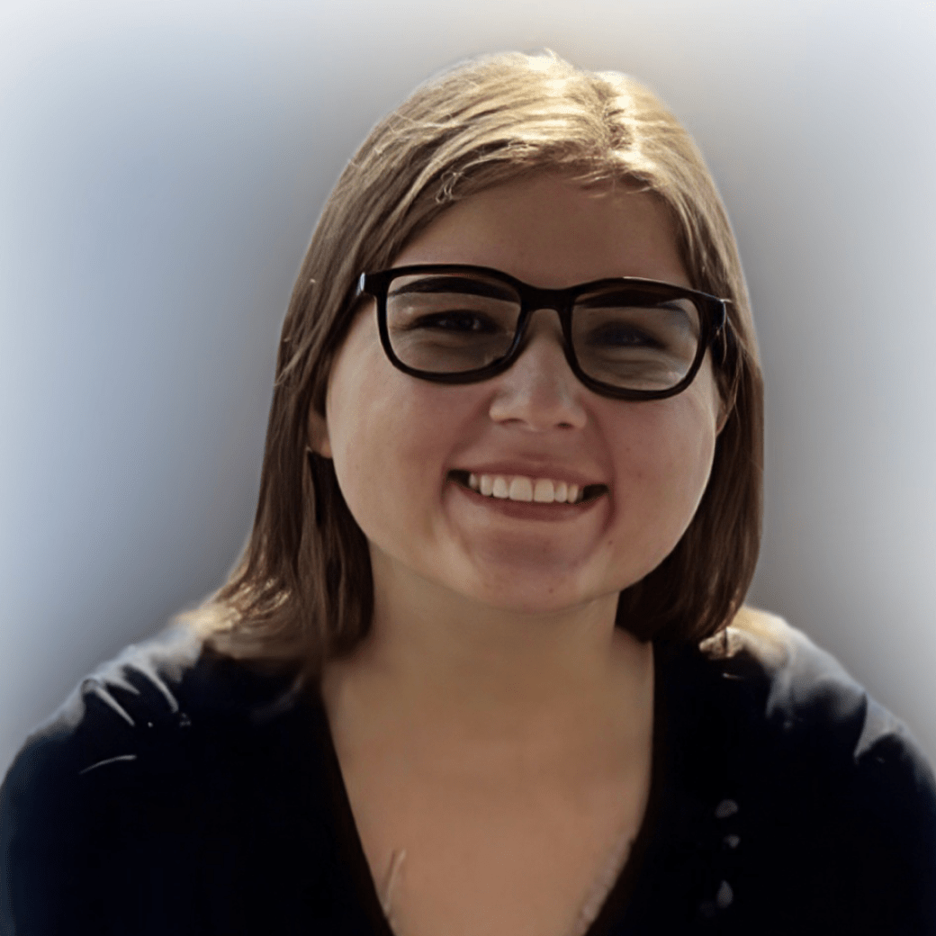 Smiling woman with short light-brown hair and black glasses, wearing a dark top, against a soft blurred background.