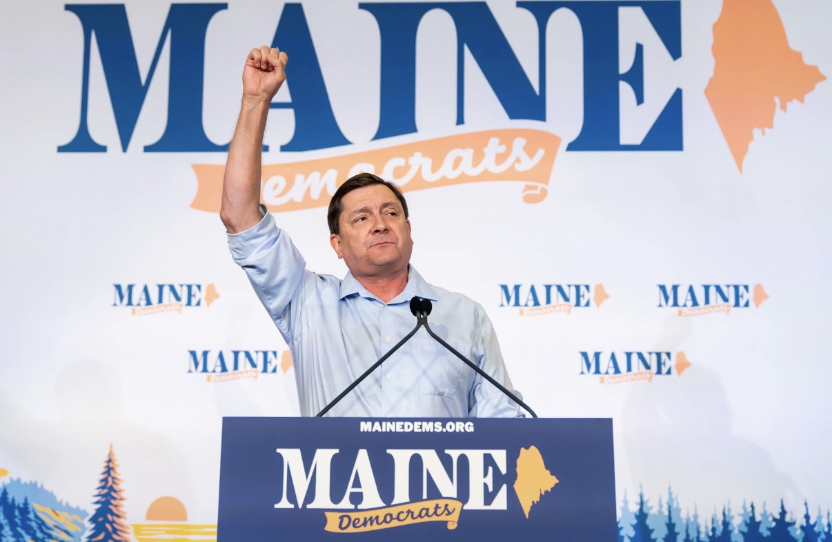 Man raising his right fist at a Maine Democrats podium during a campaign event; backdrop reads 'MAINE Democrats' with state silhouette.