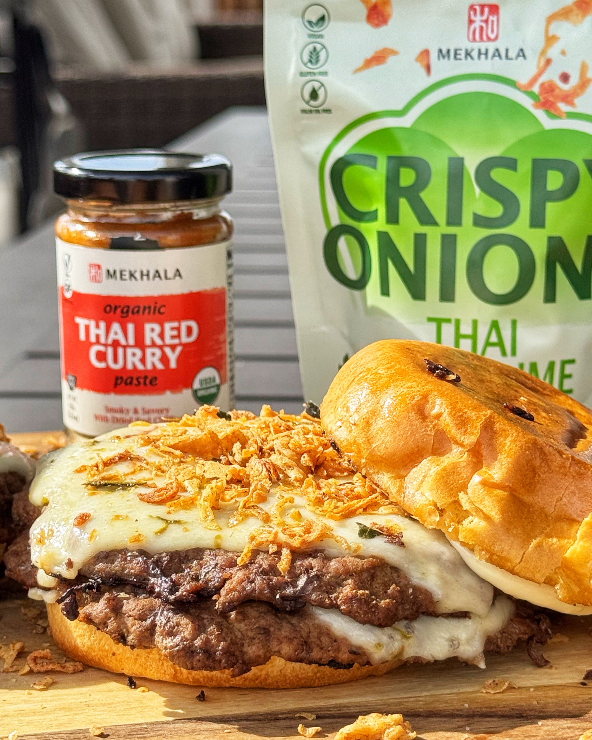 Thai Red Curry Smash Burger sliced in half showing the crust, melted cheese, and crispy onions, with Mekhala Thai red curry paste in the background