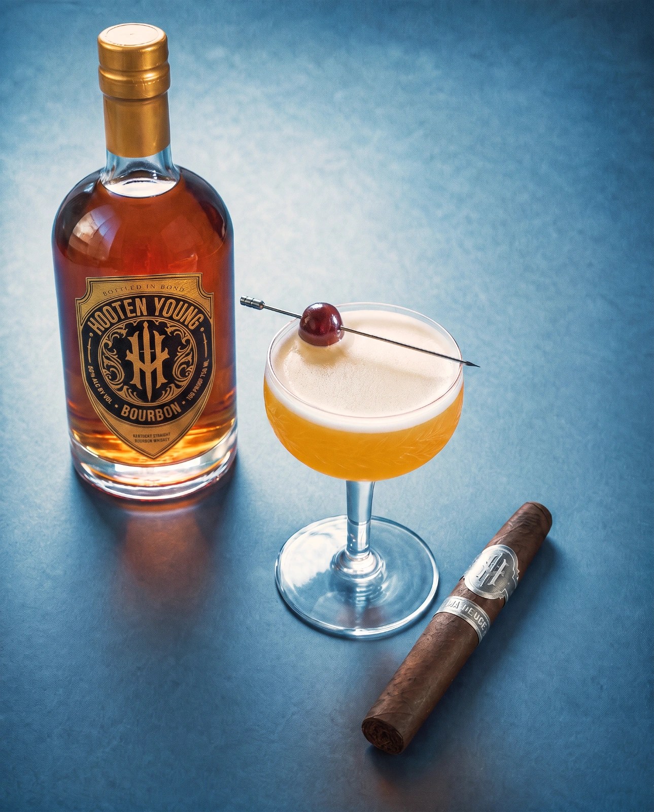 Hooten Young bourbon bottle beside a peach sour with foam and cherry garnish