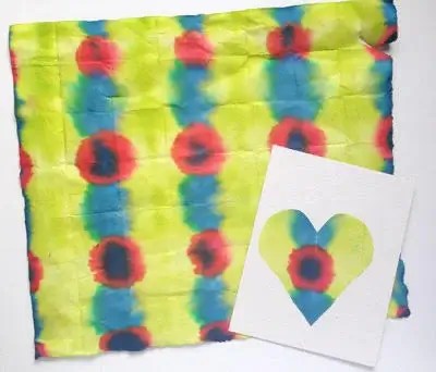 A colorful piece of Japanese tie-dyed paper featuring a repeating pattern of red, blue, and green circular designs on a yellow background. Next to it is a white card with a heart-shaped cutout, showing part of the same tie-dye pattern through the shape.
