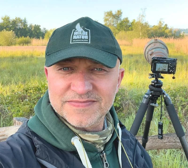 Markus mit WildeNatur-Cap vor Kamera auf Stativ in grüner Feuchtwiese.