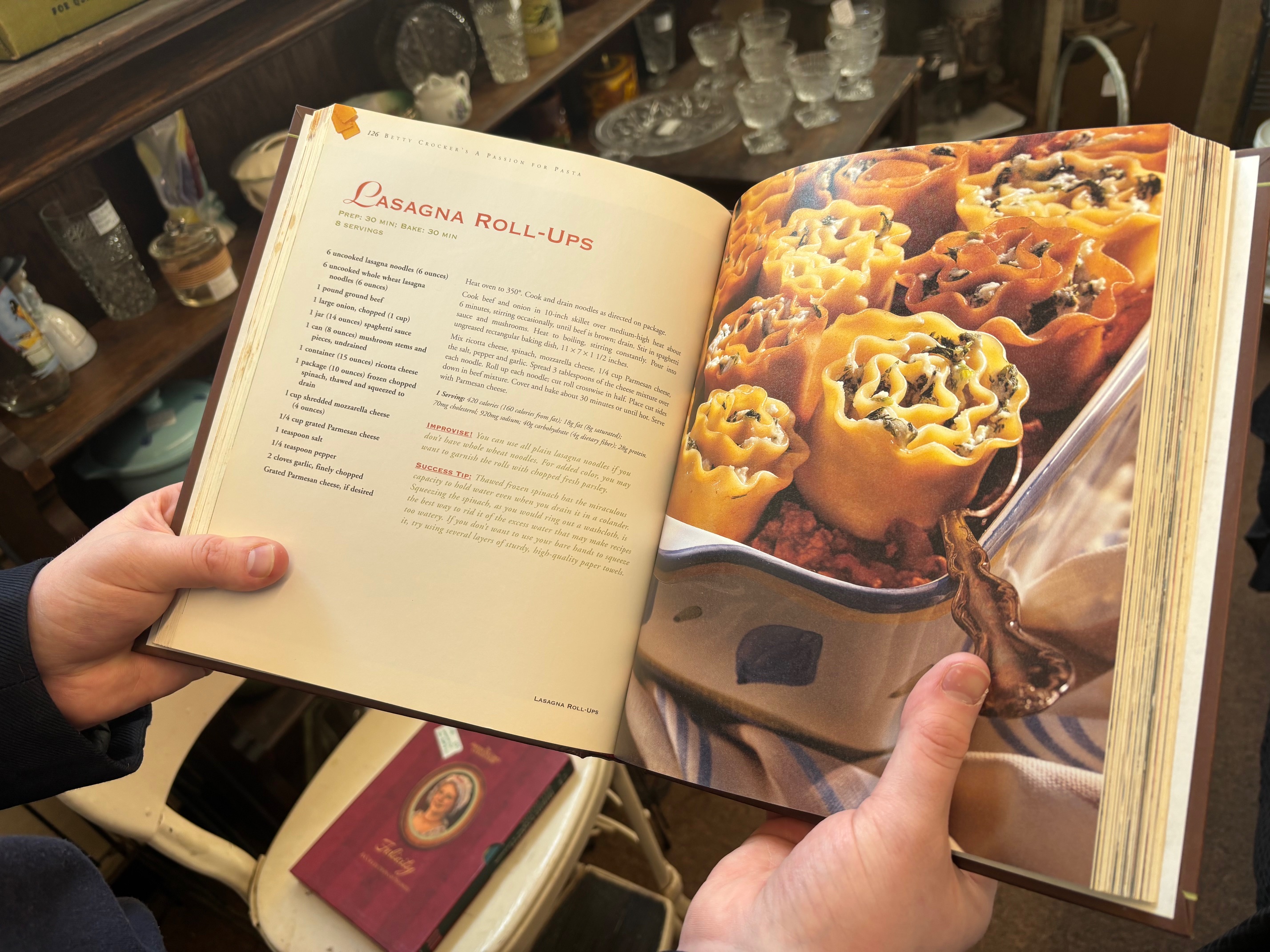 A person holding an open cookbook displaying a pasta recipe, with images of stuffed pasta dishes.
