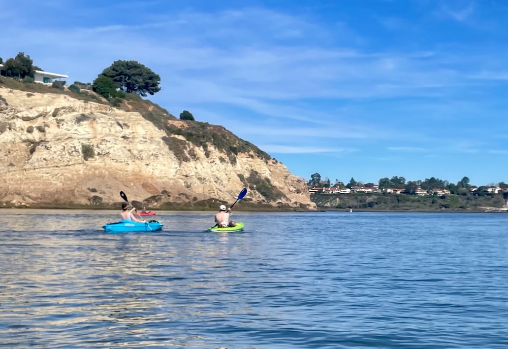 Two kayakers paddling in a placid bay, a sandstone cliff rising sharply at one side of the bay with a modern house at the top of the cliff.