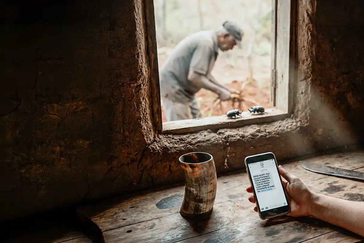 A hand holding a smartphone with a bank‑fee alert inside a dim adobe hut, a man’s blurred silhouette outside in the subtropical heat.