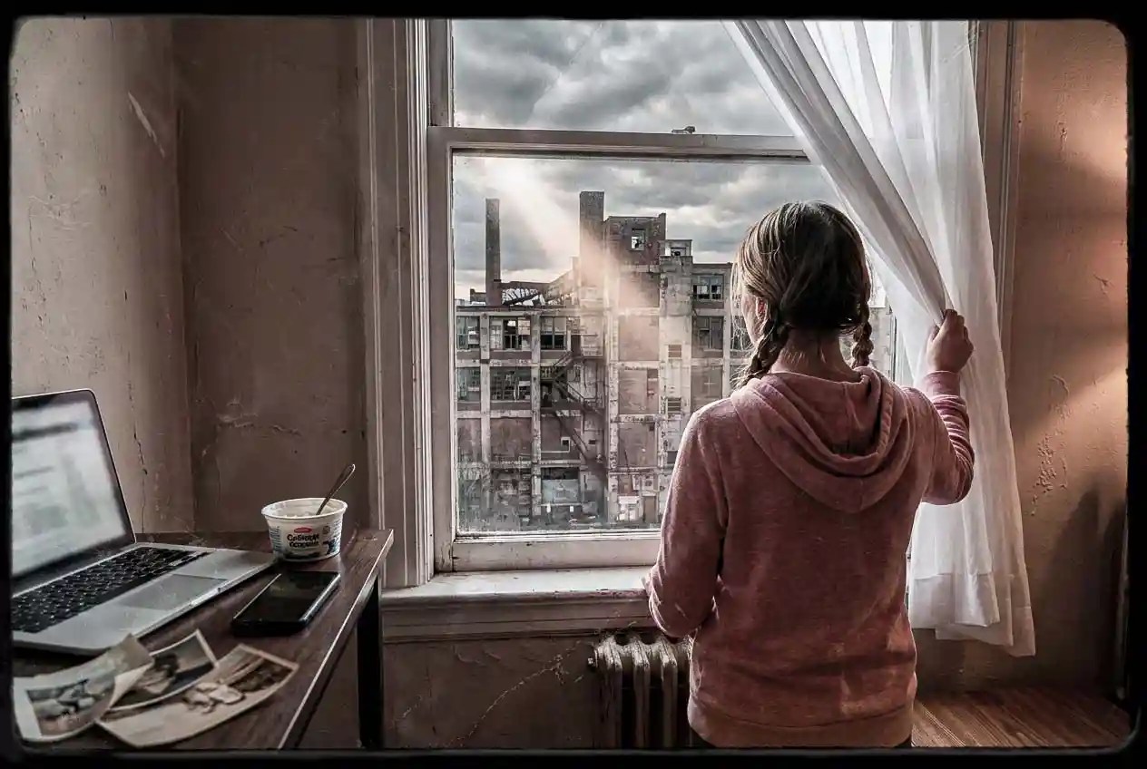 A young woman in a pink hooded top with two dark blonde braids pulls back a white curtain to look out her worn apartment window toward an old vacant factory, late afternoon light cutting through the room onto the desk with her laptop, yogurt cup, phone, and scattered photos.