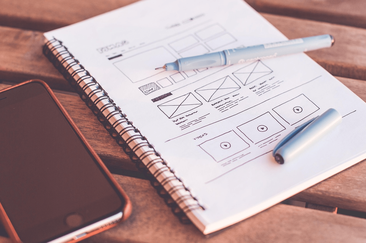 Web design planning sketch with wireframe layout on notebook beside smartphone and pen