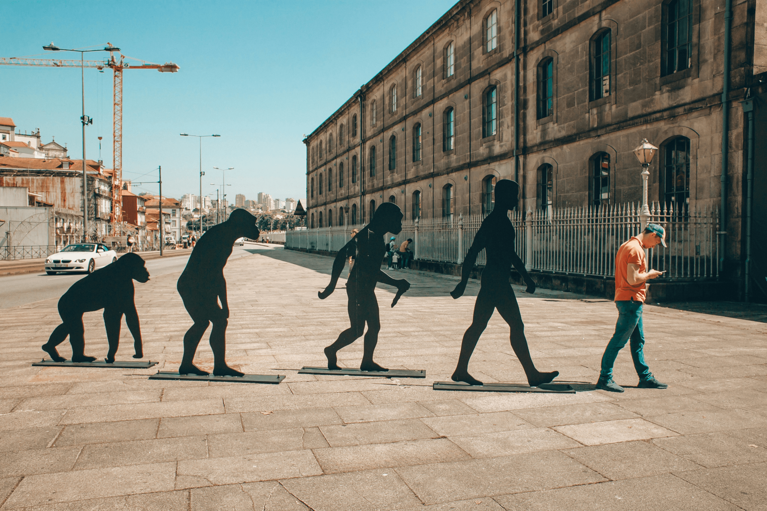 An outdoor picture of a man looking down at his smartphone in front of silhouettes of an ape evolving into a human.