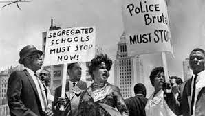 Black and white photo of civil rights protesters holding signs that read SEGREGATED SCHOOLS MUST STOP NOW! and POLICE BRUTALITY MUST STOP. The group appears to be marching in a city.