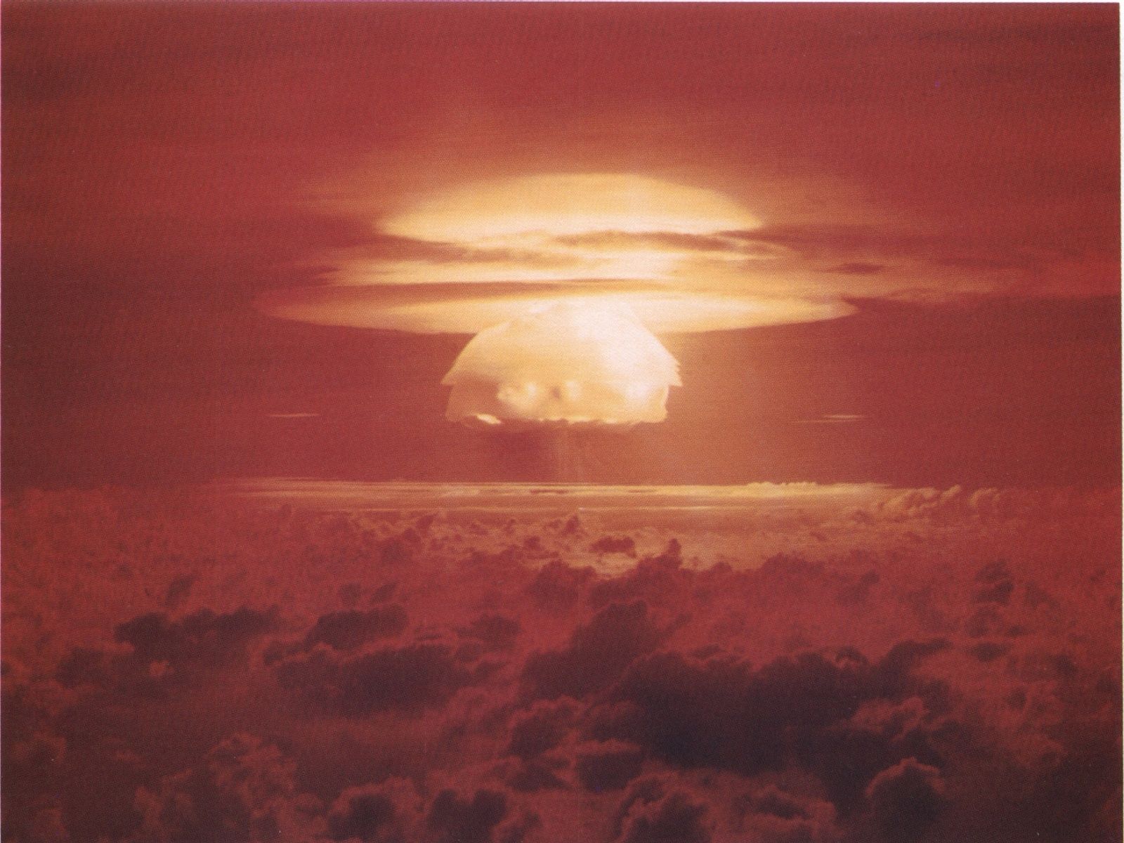 A large, bright mushroom cloud rises above dark clouds at sunset, with the sky illuminated in shades of red and orange.
