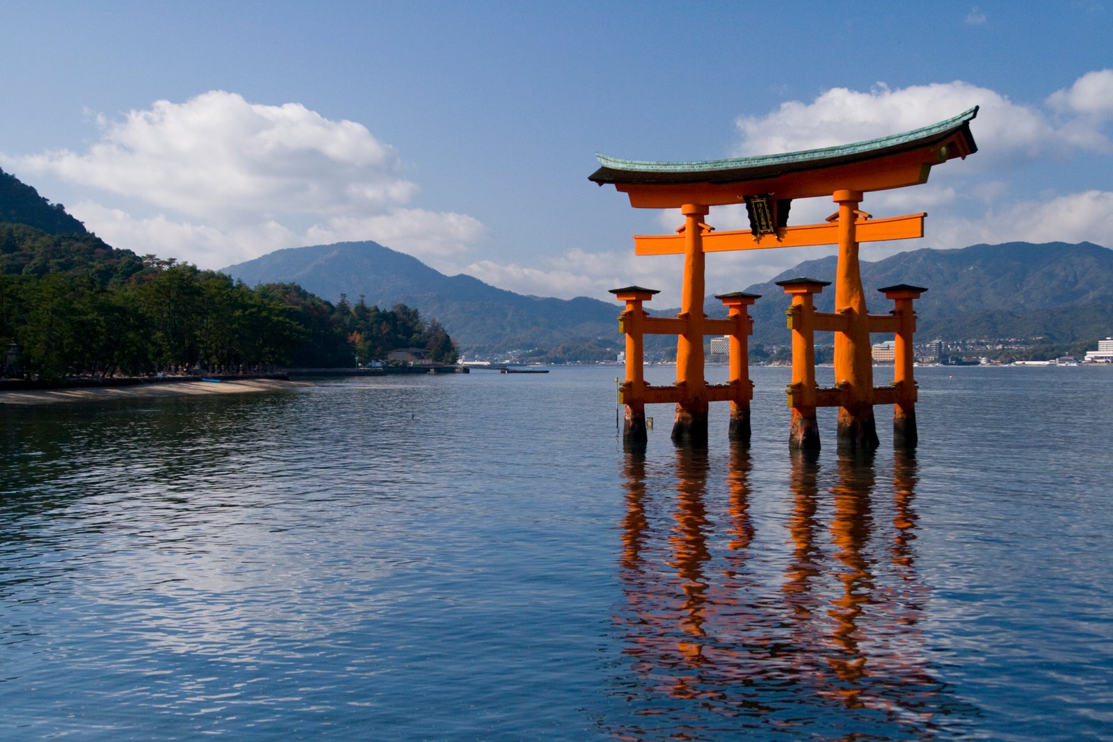 A large, traditional red torii gate stands in the water near the shore, with mountains, trees, and blue sky with clouds in the background. The gate’s reflection appears on the calm surface of the water.