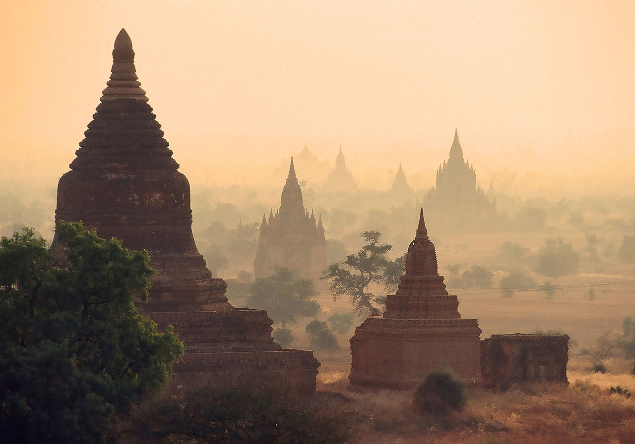 Ancient pagodas and temples in Bagan, Myanmar, rise through a golden morning mist, with trees scattered among the historic structures under a hazy sky.