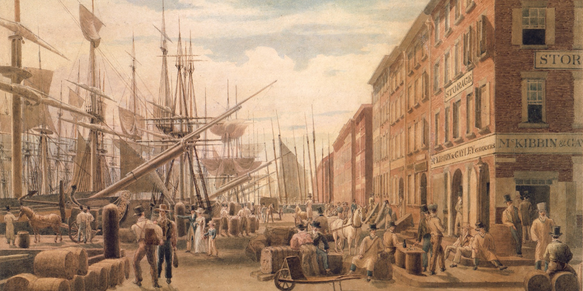 A busy 19th-century port scene shows workers loading and unloading cargo near tall sailing ships docked beside brick warehouses and storefronts, with people walking and barrels and crates scattered along the street.