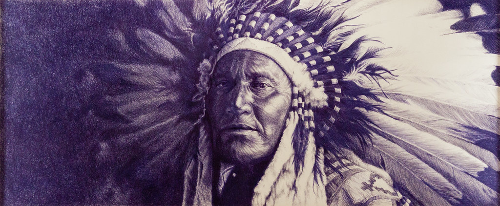A detailed, realistic drawing of a Native American man wearing a traditional feathered headdress and patterned clothing, looking solemnly ahead with dark shading and intricate textures in the feathers and face.