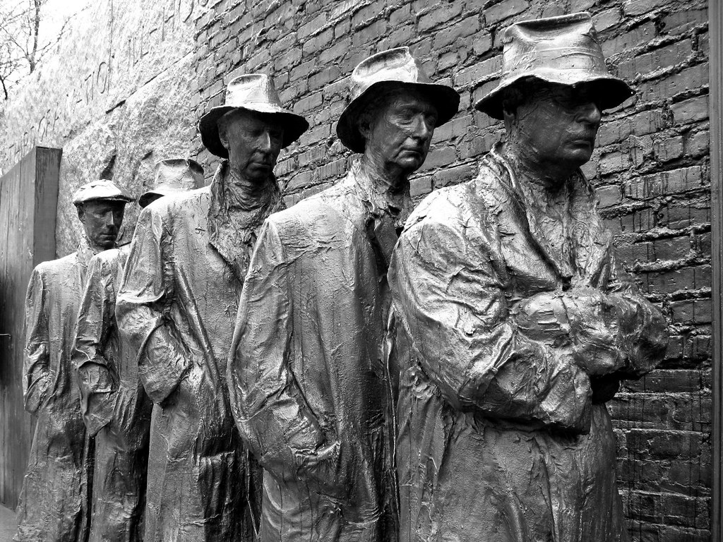 Bronze statues of men wearing coats and hats stand in a line against a brick wall, representing people waiting in a breadline during the Great Depression.