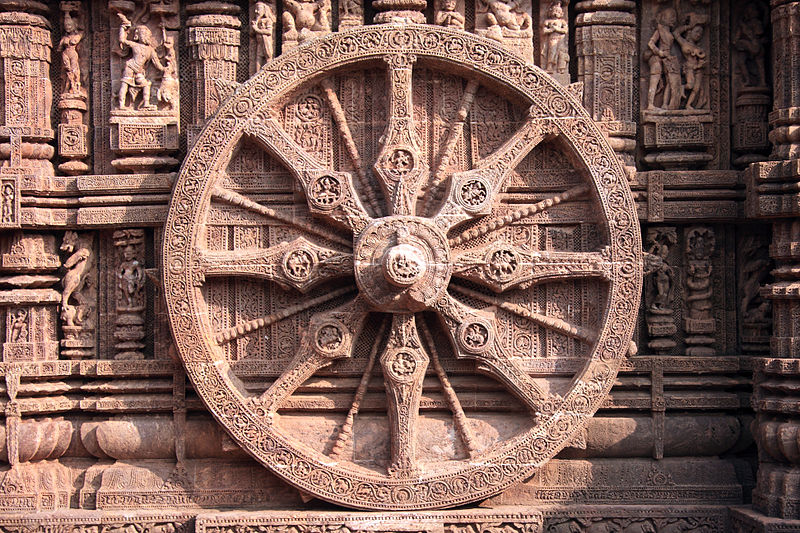 Intricately carved stone wheel with 16 spokes, part of the Sun Temple at Konark, India, set against a richly detailed wall featuring various sculptures and decorative patterns.