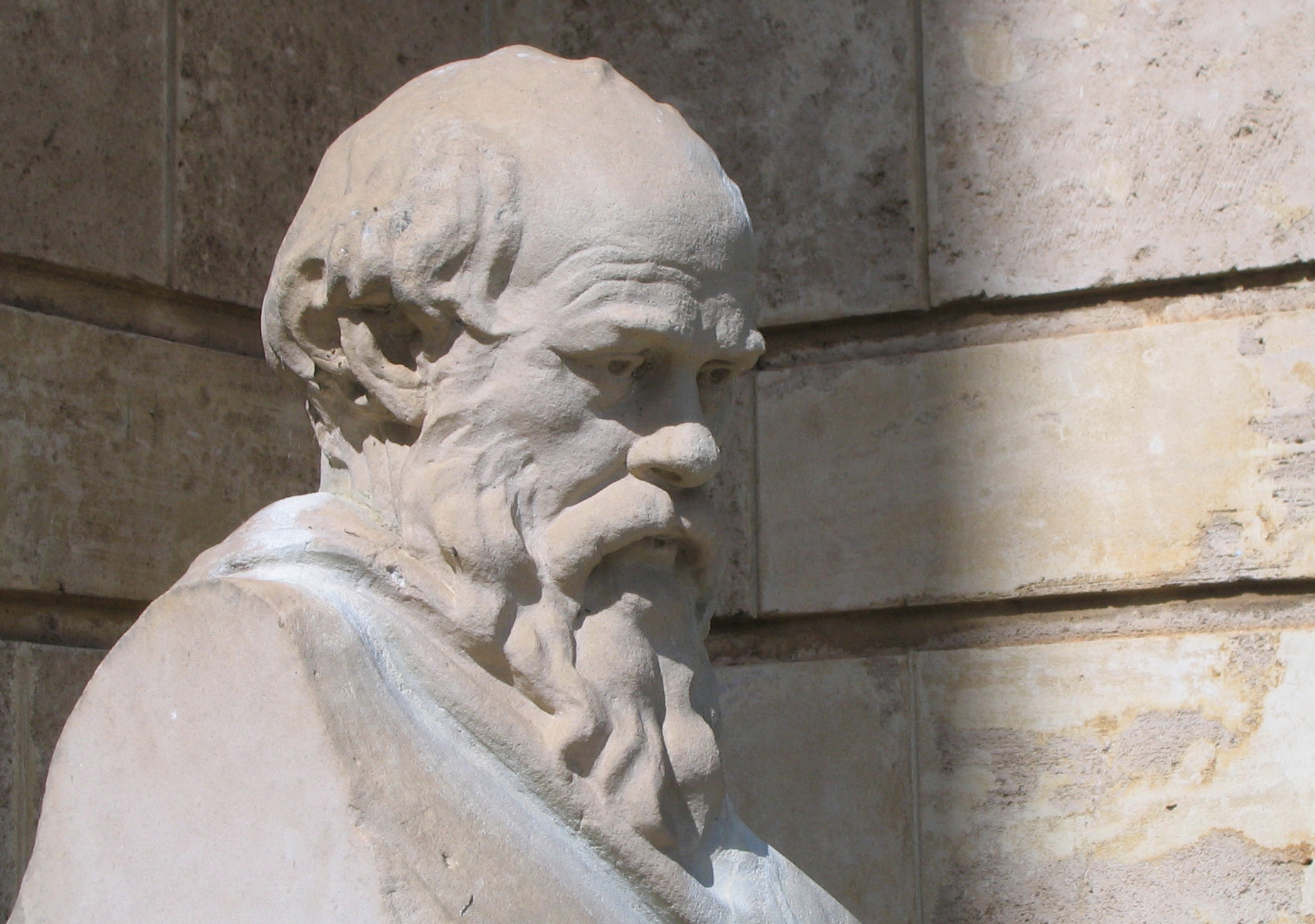 A close-up of a stone statue depicting an older bearded man with a thoughtful expression, set against a textured stone wall background.