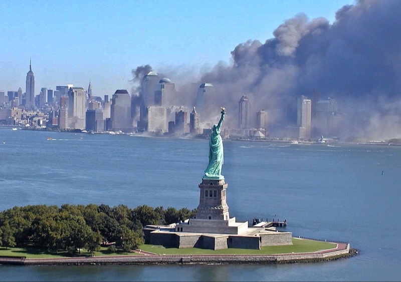 The Statue of Liberty stands in the foreground with smoke billowing from buildings in Lower Manhattan across the water, under a clear blue sky.