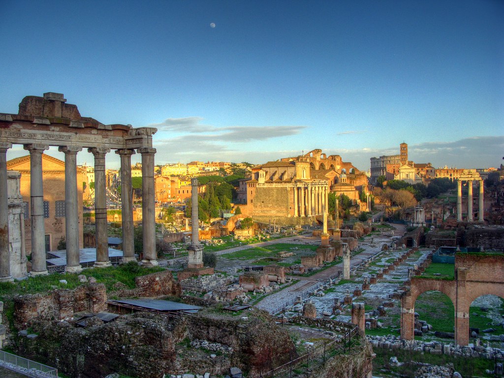 Ancient Roman ruins with tall stone columns, arches, and scattered remains, set against a cityscape under a blue sky at sunset; the scene is the Roman Forum in Rome, Italy.