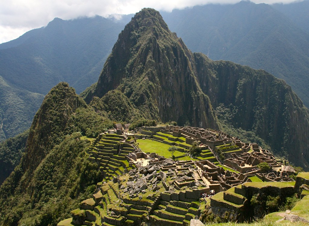 Ancient stone ruins of Machu Picchu sit atop a lush, green mountain ridge with steep terraces and pathways, surrounded by tall, forested peaks under a partly cloudy sky in the background.