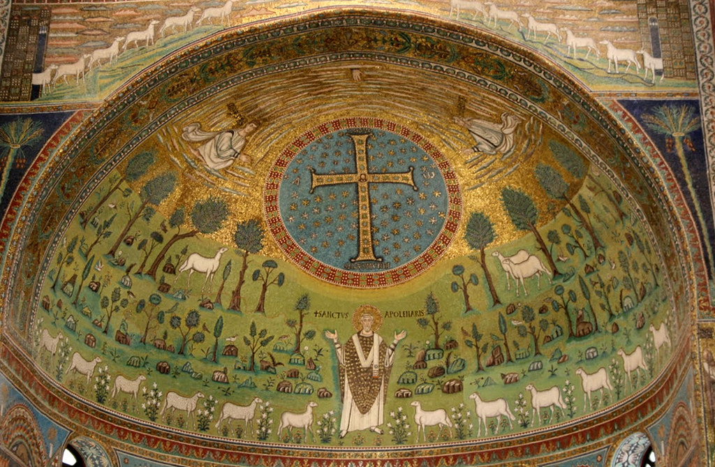 A detailed mosaic featuring a large cross surrounded by gold and blue, sheep grazing among trees, and a robed figure with outstretched arms, with angels above, set in a richly decorated arch.