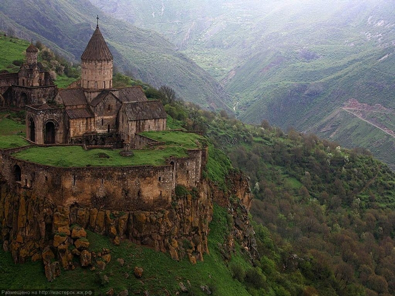 An ancient stone monastery sits atop a rocky hill, surrounded by lush green mountains and valleys, with mist in the background.