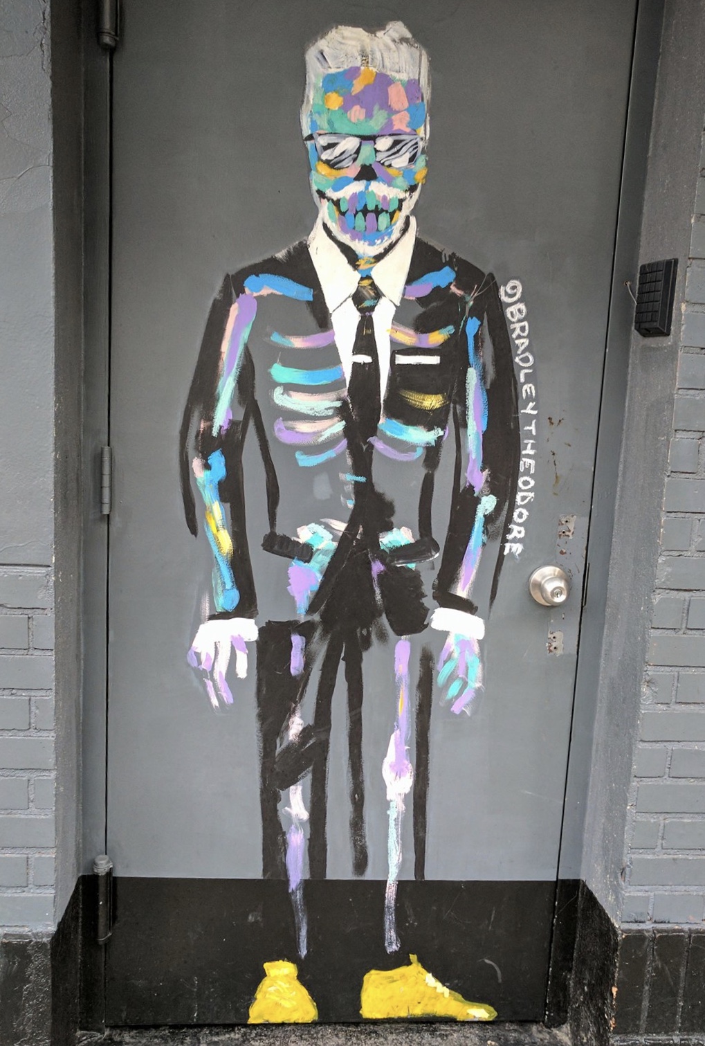 A Bradly Theodore skeleton in bold colors wearing a dark suit and tie, with yellow shoes and sunglasses. Street art that survived.