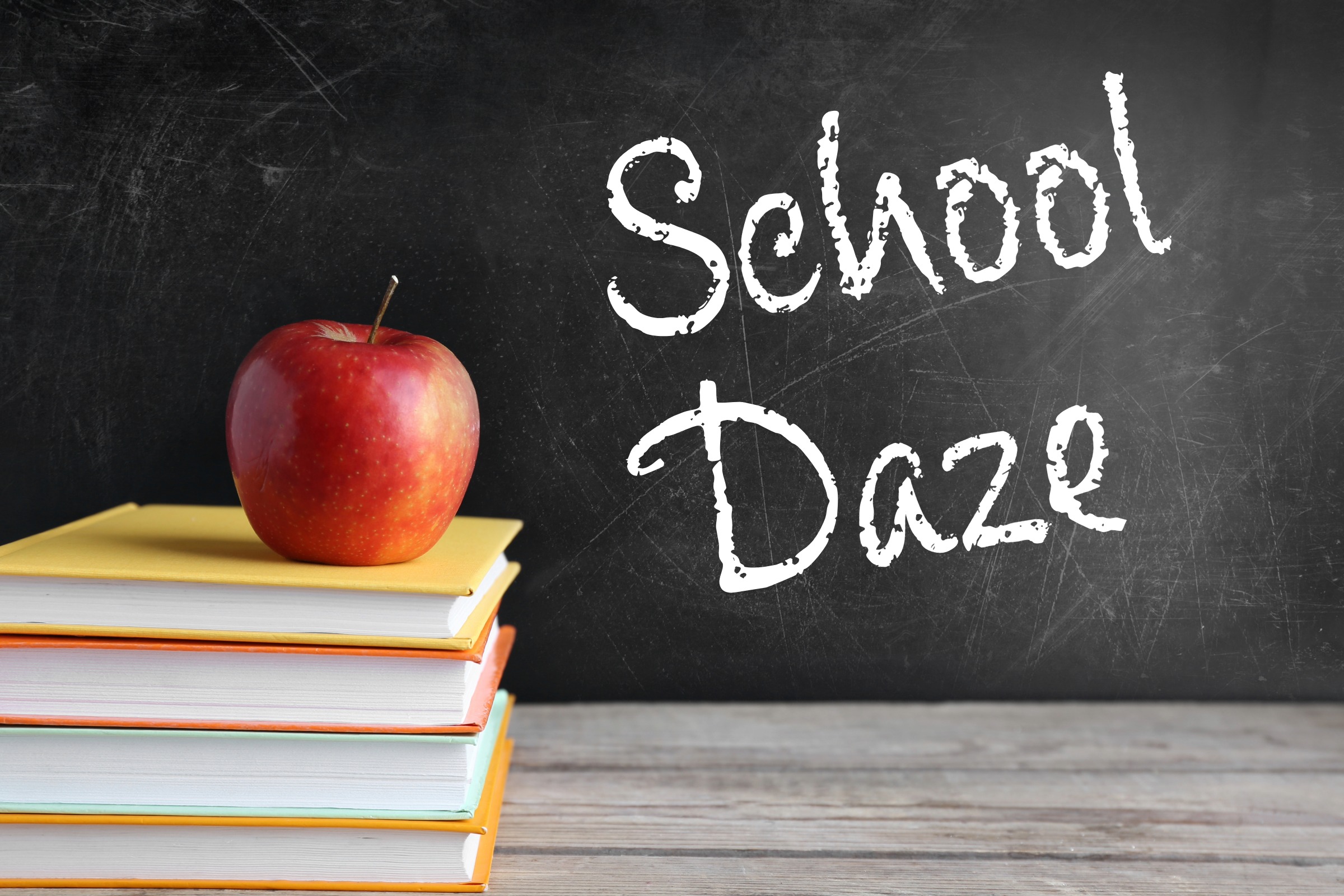 A stack of books with an apple on top with a blackboard in the background that has "School Daze" written on it in chalk.
