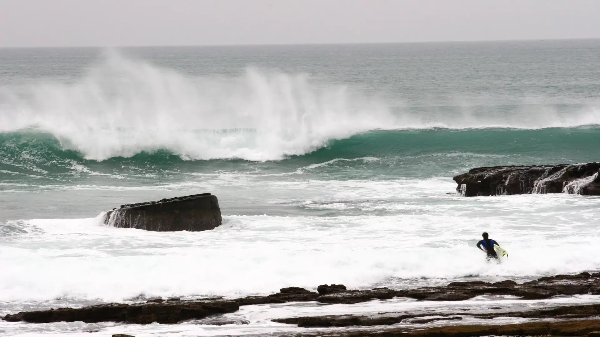 A surfer in a wetsuit stands on rocky shore with a surfboard, gazing at large crashing waves in an overcast ocean—an inspiring scene near surf school Taghazout, home to Sunset Surfhouse and unforgettable surf lessons Morocco offers.
