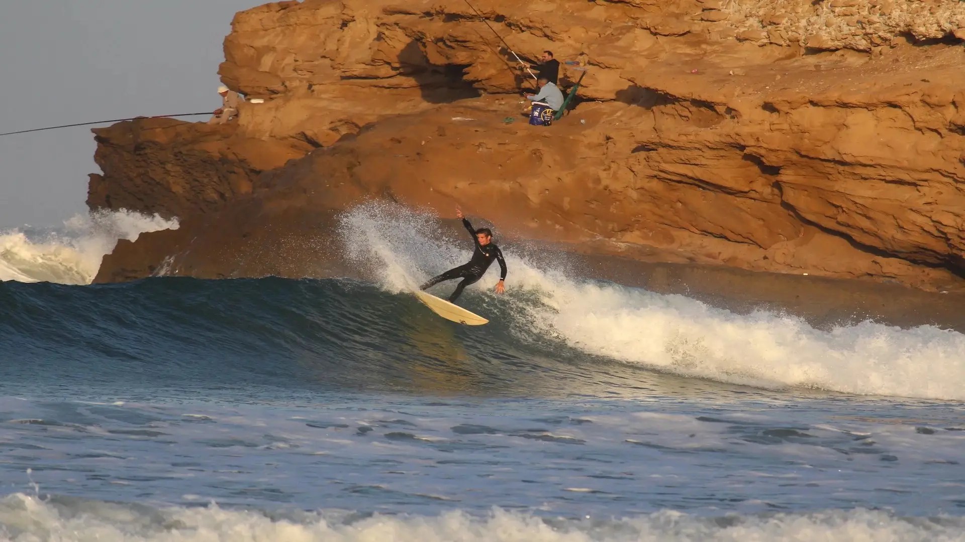 A surfer in a black wetsuit rides a wave near a rocky shoreline, kicking up spray as they turn—a classic scene for Morocco surfing enthusiasts. In the background, two people sit on the rocks, one holding a fishing rod.