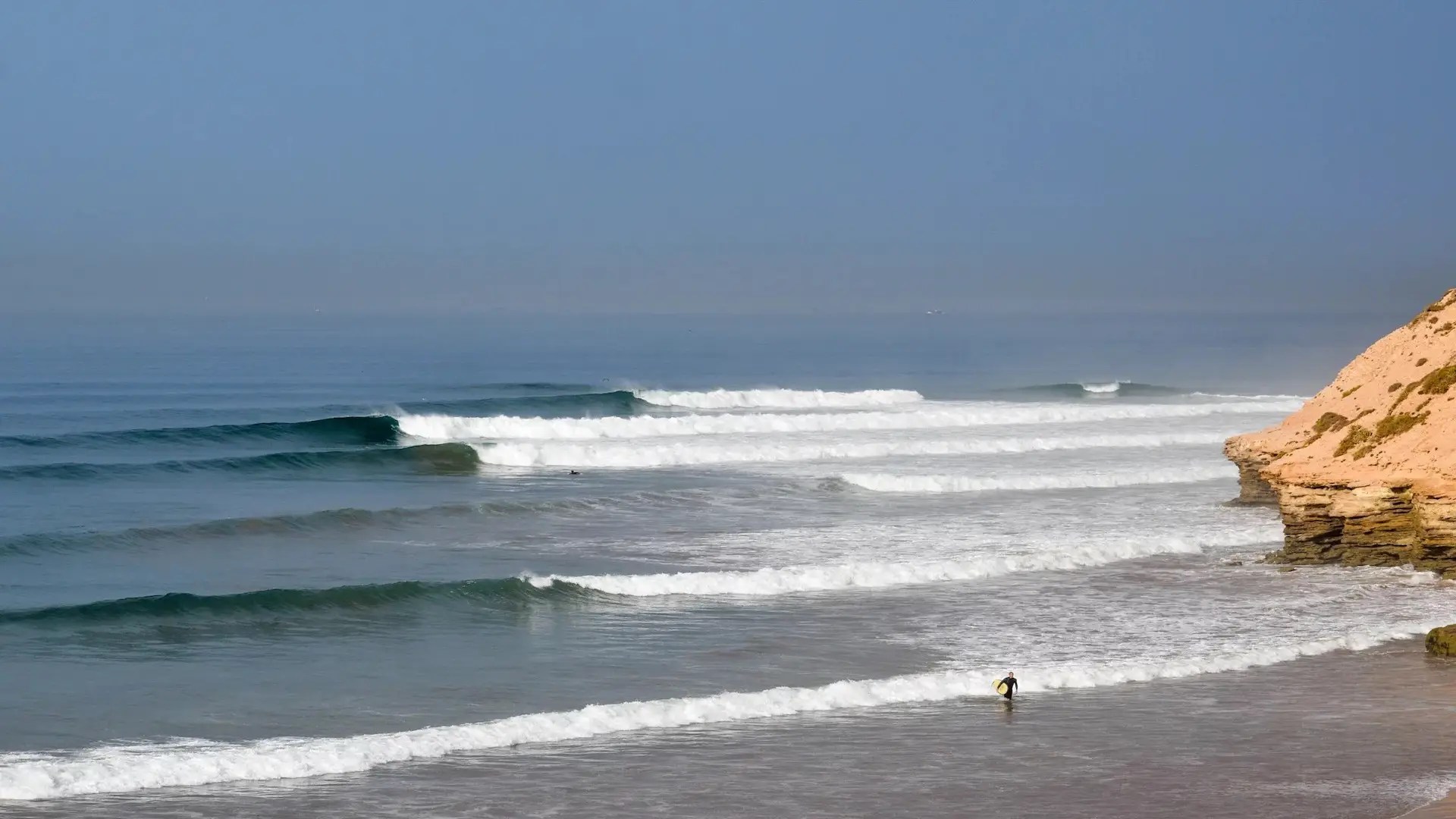 A sandy beach with waves rolling in from the blue ocean; a lone person carrying a surfboard walks along the shore near rocky cliffs under a clear sky, perfect for beginner surf lessons or joining a surf camp Tamraght.
