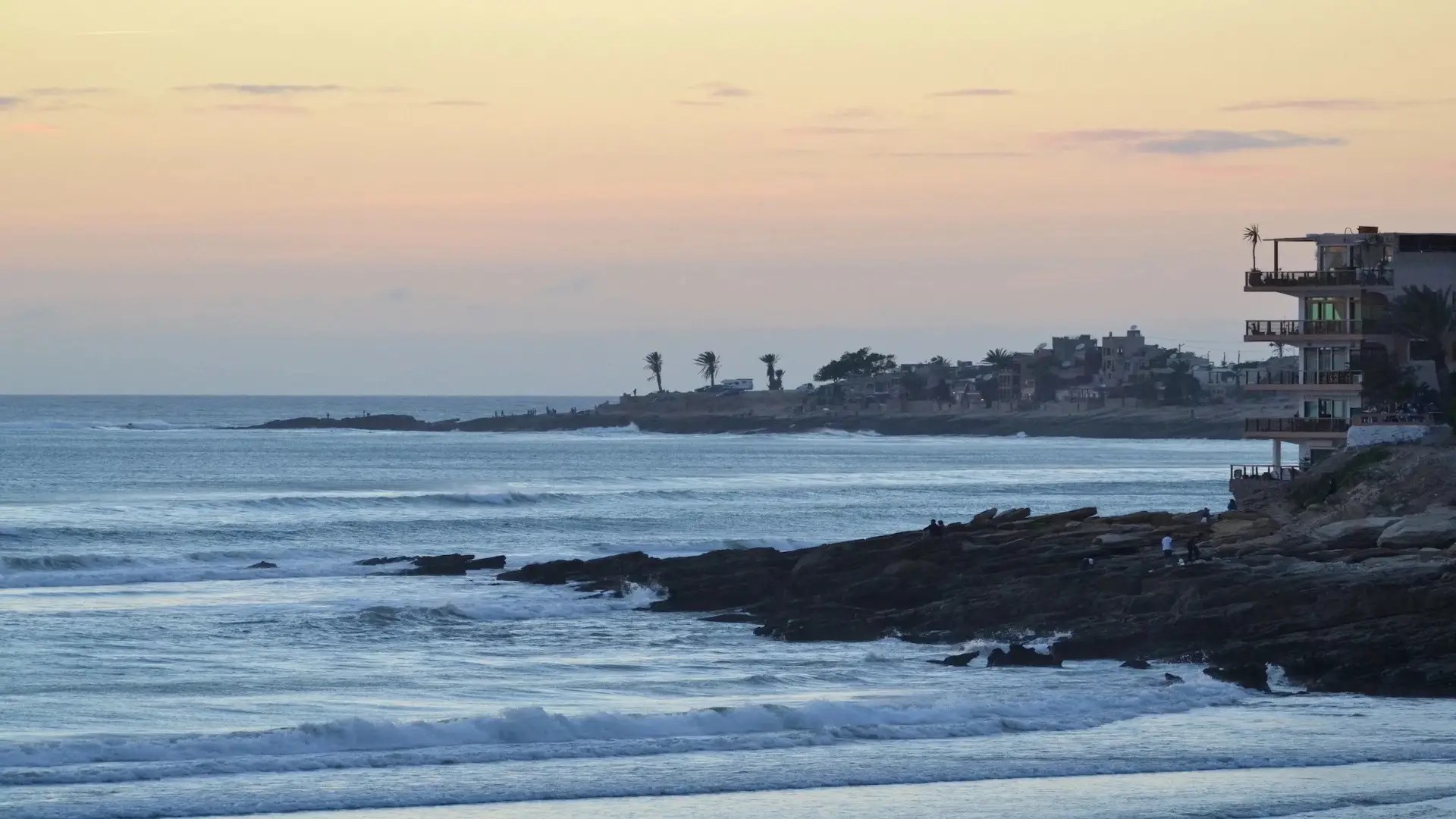 Calm ocean waves roll onto rocky shores at sunset, pastel orange and purple hues in the sky. In the distance, a coastal town known for Morocco surfing and beginner surf lessons lines the shoreline amid palm trees and scattered buildings.