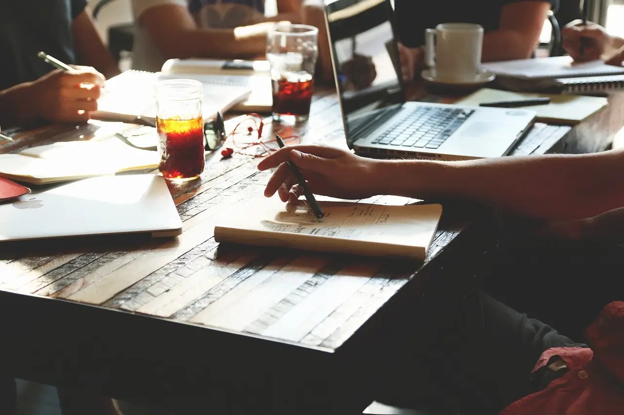 People sitting around a table doing a group work. There are notebooks, glasses with soda and a laptop on the table.