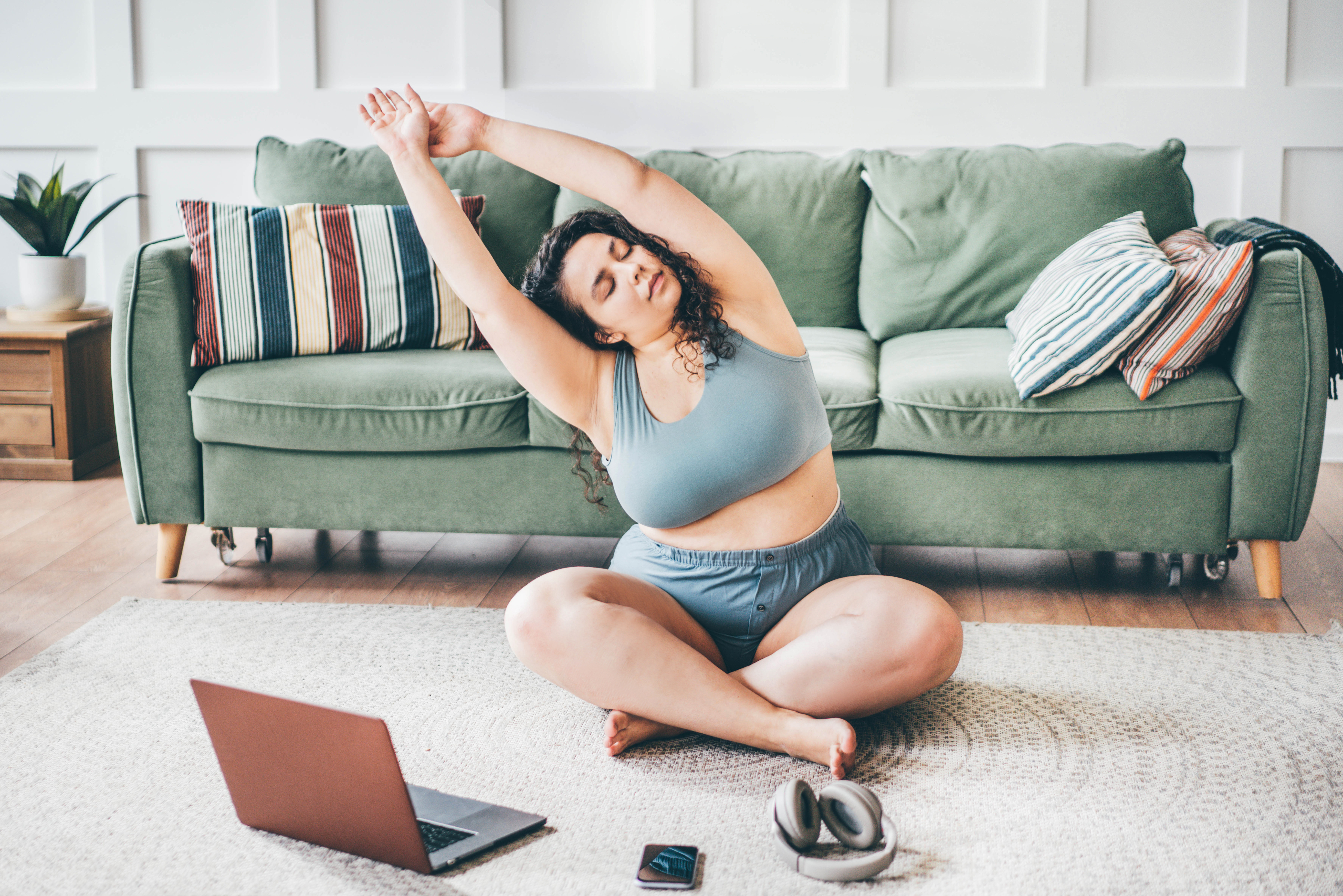 How can I lose weight? Curly haired overweight young woman in top and shorts turns on online yoga training and practices exercises on floor mat against green sofa by wall