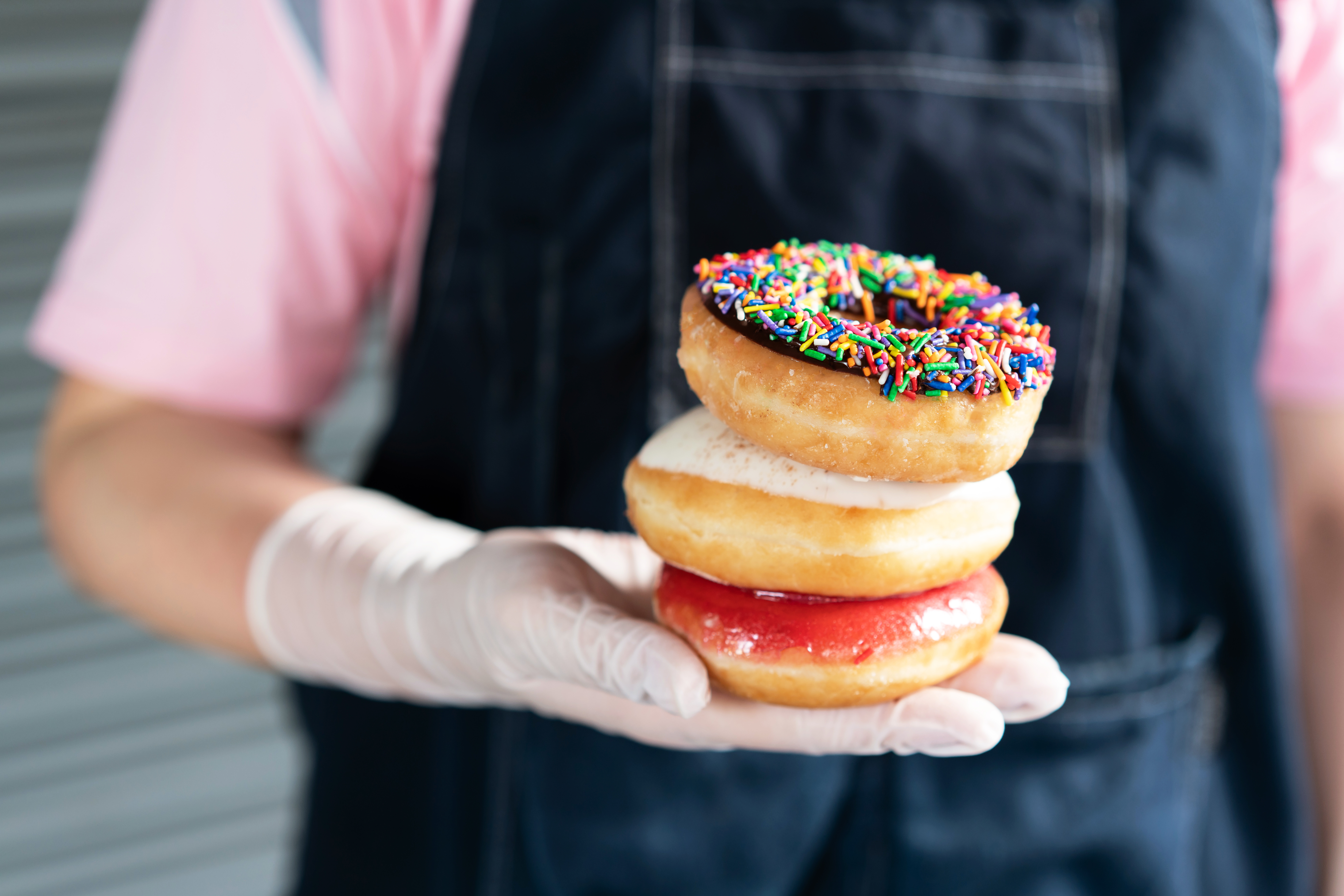 Trans Fat. Stacking chocolate frosted donut with sprinkles, sugar-glazed frosted and Bavarian donut on woman hand that wearing latex gloves. Playful and joyful tasty sugary comfort food for customers.