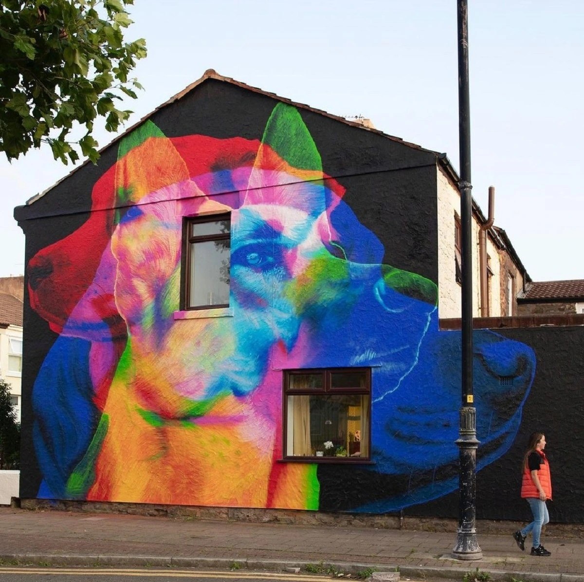 The gable of a house in New Brighton painted with three overlapping dog portraits in red, rainbow colors and blue, with a woman walking past on the sidewalk.