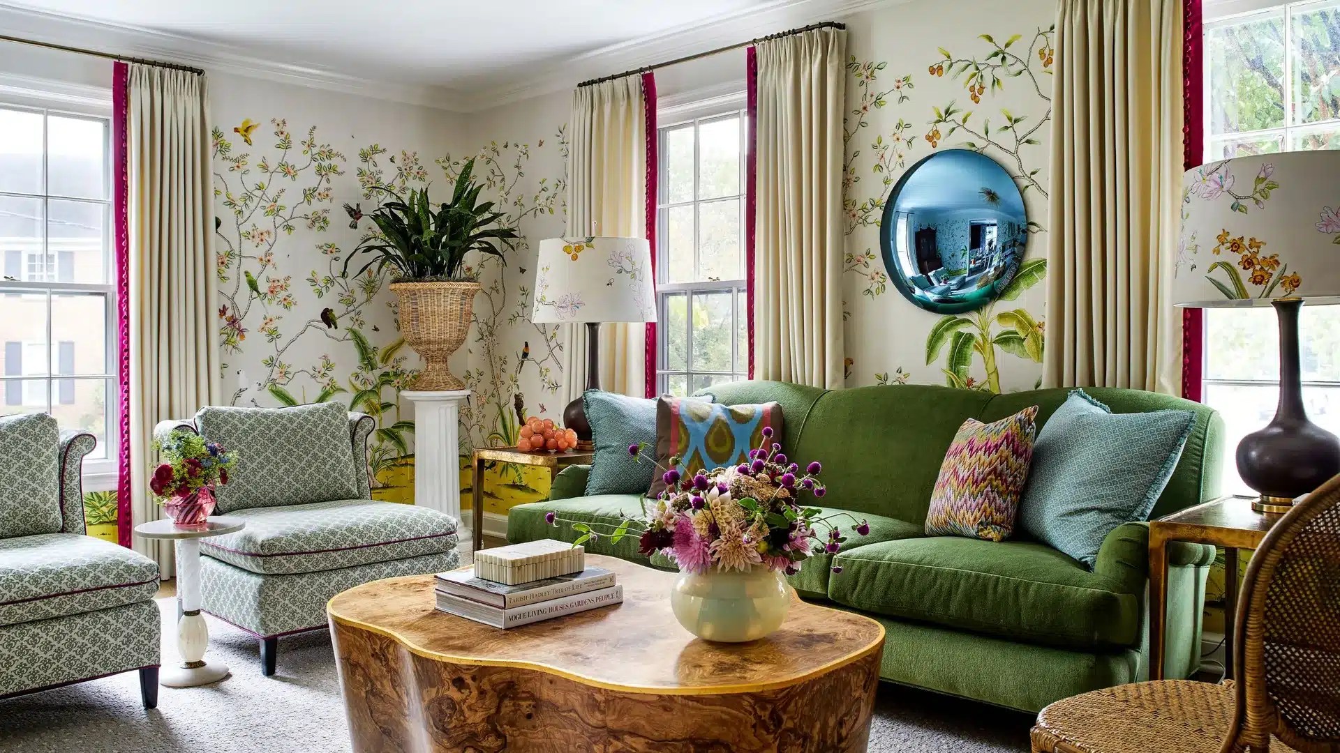 A colorful living room featuring scenic wallpaper, a green sofa, patterned armchairs, vintage artwork and layered decorative accessories inside a beautifully renovated home.