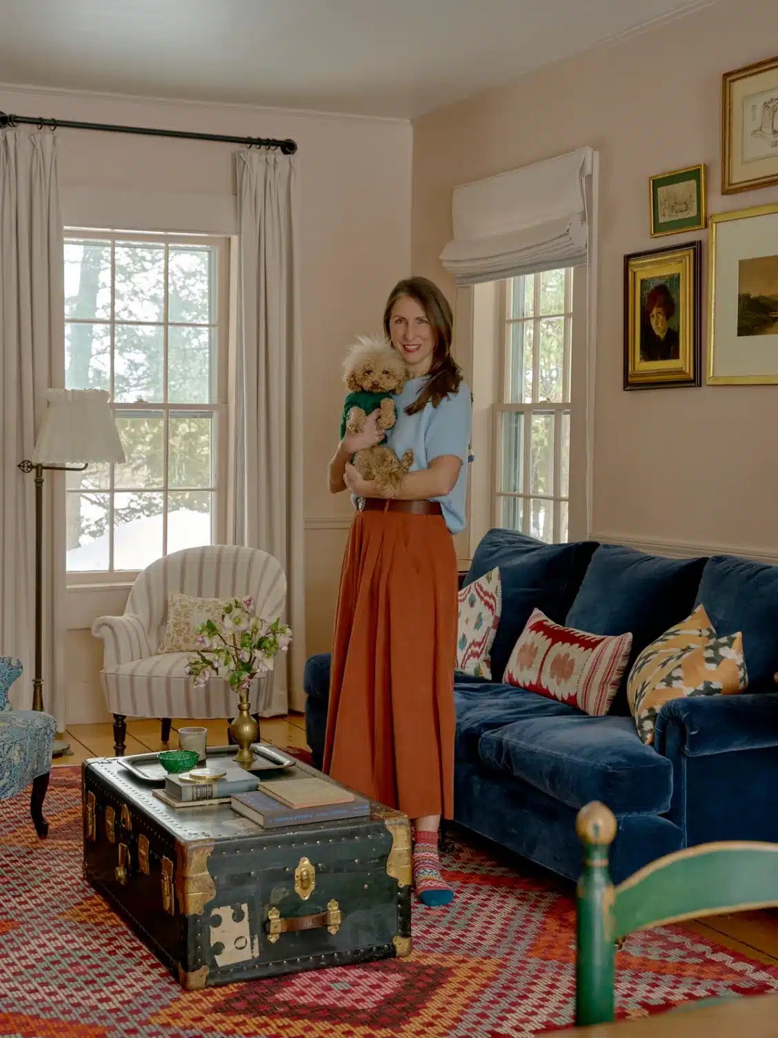 A colorful contemporary living room featuring a curved rust-colored sofa, abstract artwork, colorful accent chairs, a patterned area rug, sculptural lighting and layered decorative accessories that create a warm, modern interior.