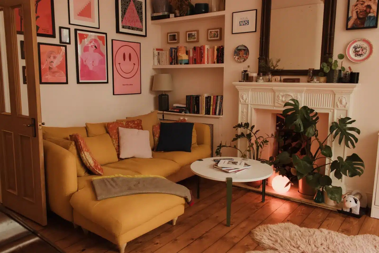 A colorful living room with blush pink painted walls, a mustard yellow sofa, framed gallery wall, built-in shelving and period architectural details in a London apartment.