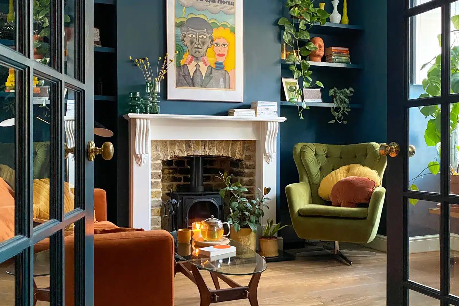 A colorful living room featuring deep teal walls, a burnt orange sofa, green armchair, fireplace, patterned textiles and vintage decor inside a renovated Edwardian home.