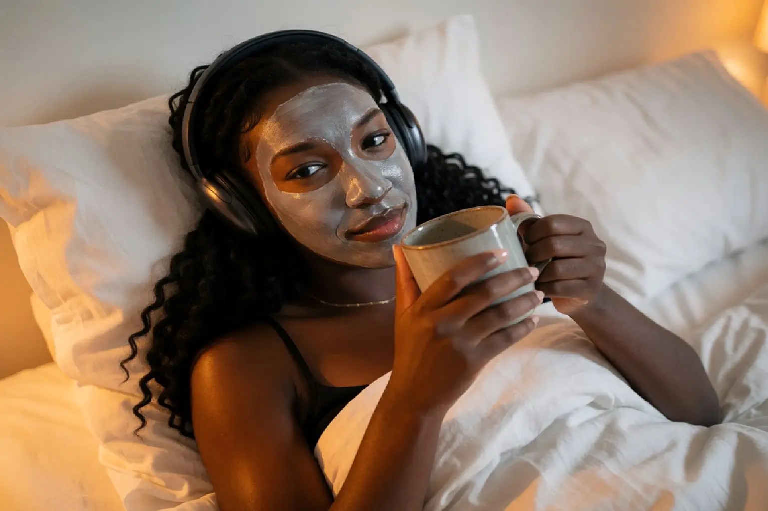 Woman relaxing in bed with a face mask, headphones and a warm drink, representing Self-Care & Wellness and everyday mental wellbeing routines.