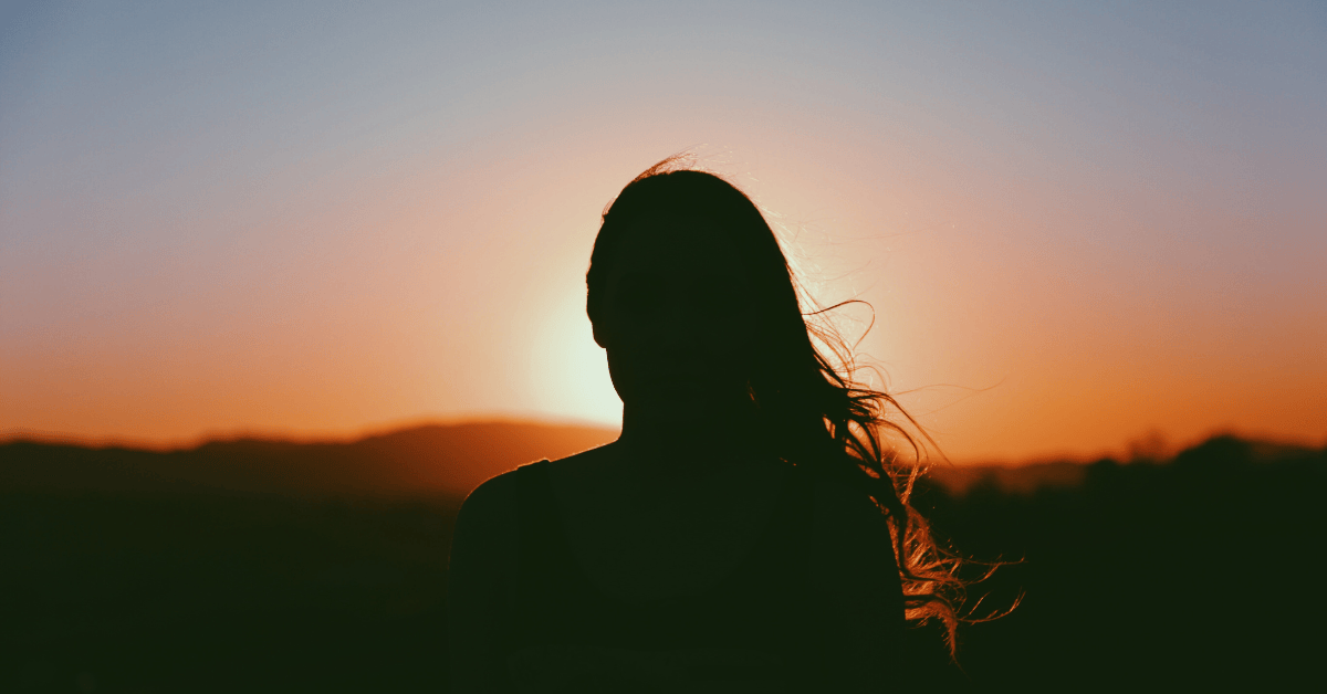 Silhouette of a woman at sunset with wind in her hair, symbolizing surrender, stillness, and spiritual transformation.