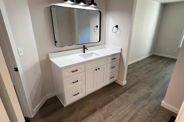 Modern farmhouse bathroom vanity with white custom cabinets, quartz countertop, black hardware, and matte black faucet under industrial-style triple light fixture and large framed mirror. Features wood-look vinyl plank flooring and neutral beige walls for a clean, contemporary home design.