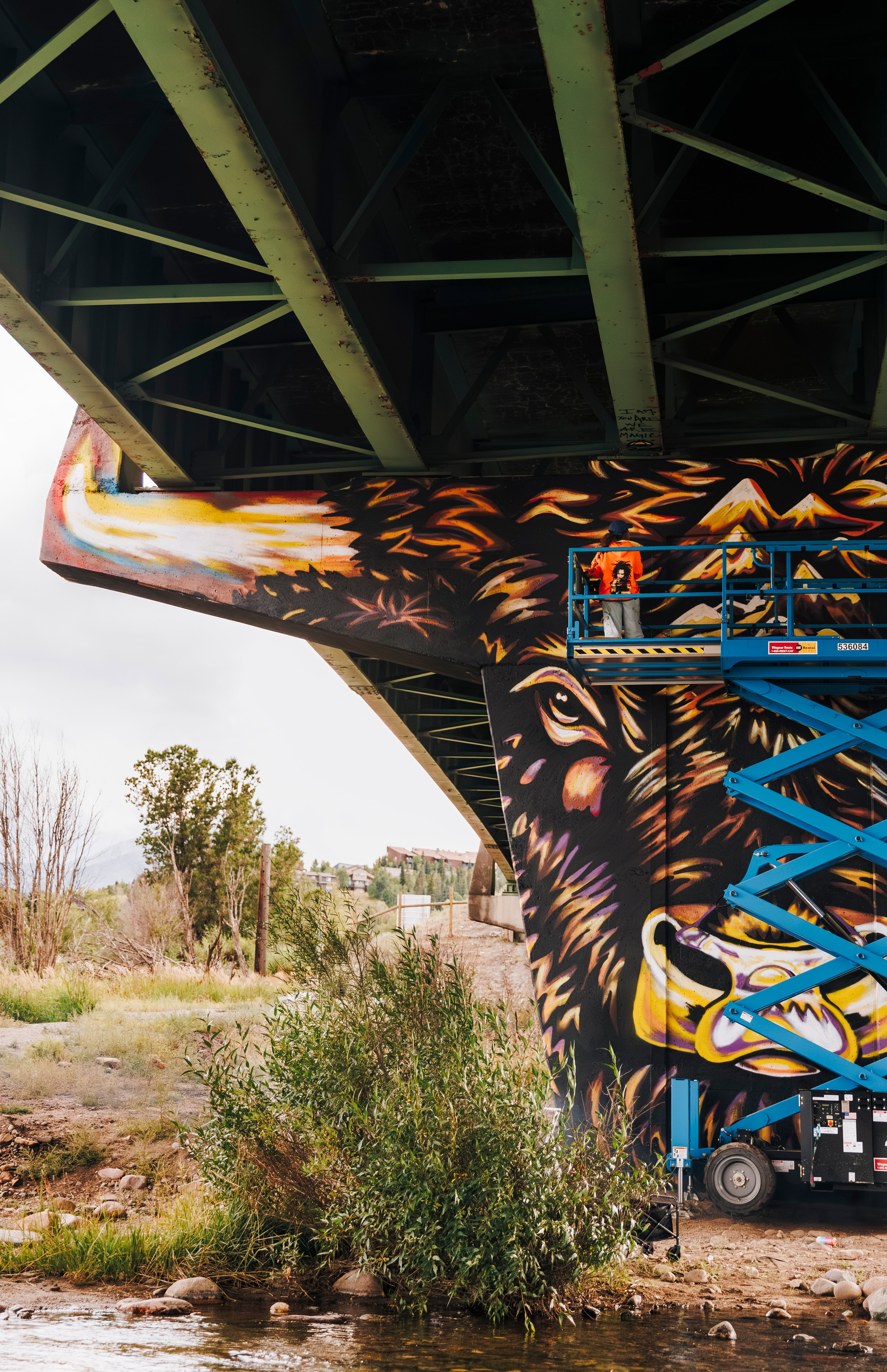 Vibrant street art mural of a bear under a bridge, painted with colorful flames and mountain scenery, using a scissor lift for precise mural work, showcasing urban art and graffiti creativity.