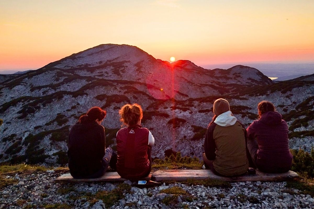 Four women sitting on a mountain bench at sunset, two on each side with a gap between them, illustrating space and independence in travel relationships.