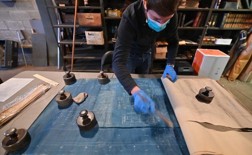 Man wearing a mask and gloves holds a tool to clean off a blue drawing.