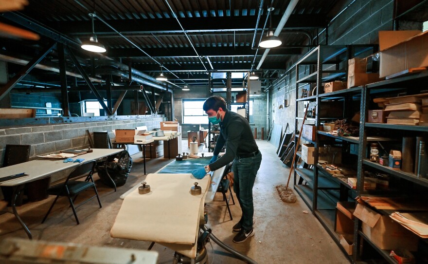 A man wearing a mask and gloves dusts off drawings on a table in the middle of a warehouse full of records.